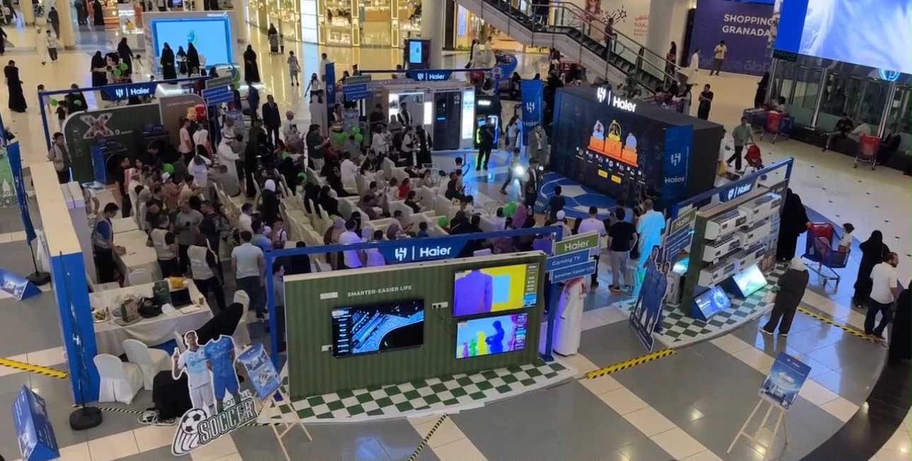 Brand activation in a shopping mall atrium, seen from above, with a seated audience, a stage, screens and product displays